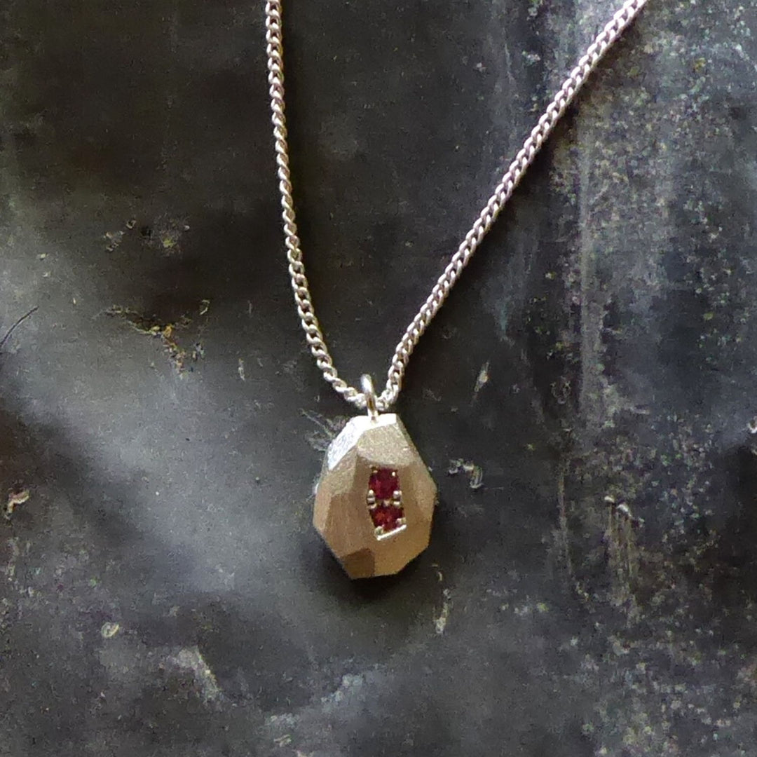 ecken-und-kanten-kette-mit-einem-kleinen-tropfenfoermigen-anhaenger-in-der-groesseren-variante-mit-unregelmaessigen-facetten-die-kette-besteht-aus-recycling-sterling-silber-bei-diesem-anhaenger-sind-auf-der-hauptfacette-zwei-rubine-eingefasst-als-verschluss-der-kette-ist-ein-karabiner-befestigt-den-man-auf-zwei-verschiedene-laengen-einhaken-kann-nachhaltiger-schmuck-von-hand-gefertigt-in-unserer-goldschmiede-in-frankfurt-bockenheim