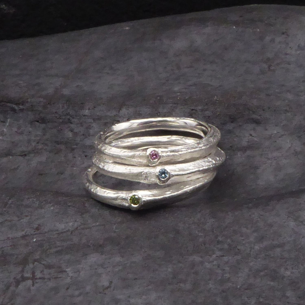 drei-aufeinander-gestapelte-astringe-mit-farbigen-brillanten-der-unterste-ring-hat-einen-grünen-dermittlere-ring-einen-blauen-und-der-oberste-einen-pinken-stein-im-blattansatz-eingefasst