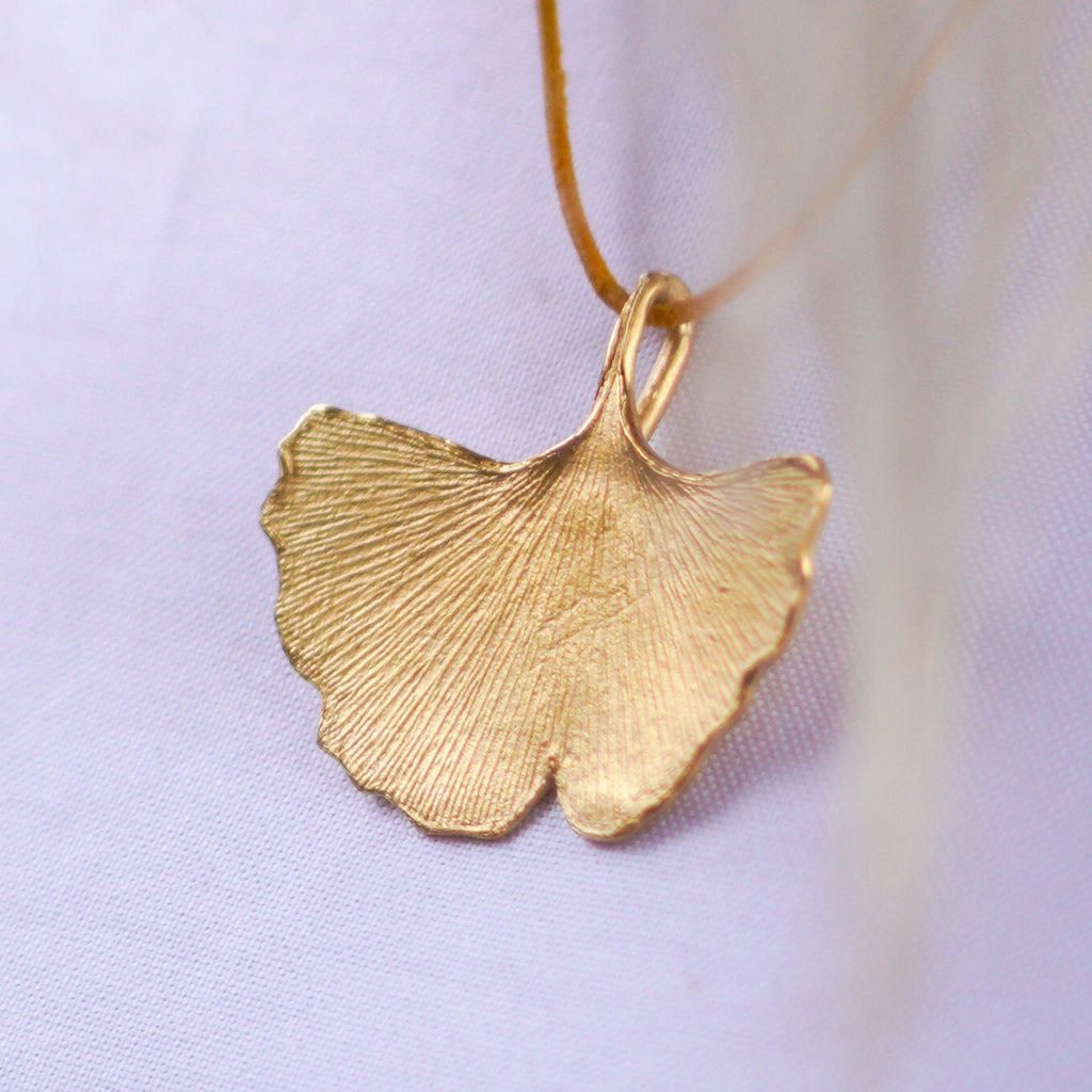 großer-handgefertigter-ginkgo-anhänger-mit-feiner-struktur-von-echten-ginkgoblaettern-abgeformt-ohne-kette-an-lederband-auf-weißem-oberteil-in-sterling-silber-vergoldet-oder-echtgold-aus-unserer-goldschmiede-in-frankfurt-auch-in-fairtrade-gold-erhaeltlich-ausladende-form-mit-einem-angedeuteten-schlitz-und-wellen-an-den-blattraendern-ungefaehr-49-millimeter