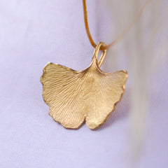 großer-handgefertigter-ginkgo-anhänger-mit-feiner-struktur-von-echten-ginkgoblaettern-abgeformt-ohne-kette-an-lederband-auf-weißem-oberteil-in-sterling-silber-vergoldet-oder-echtgold-aus-unserer-goldschmiede-in-frankfurt-auch-in-fairtrade-gold-erhaeltlich-ausladende-form-mit-einem-angedeuteten-schlitz-und-wellen-an-den-blattraendern-ungefaehr-49-millimeter
