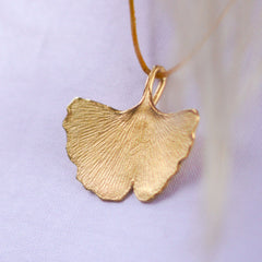 großer-handgefertigter-ginkgo-anhänger-mit-feiner-struktur-von-echten-ginkgoblaettern-abgeformt-ohne-kette-an-lederband-auf-weißem-oberteil-in-sterling-silber-vergoldet-oder-echtgold-aus-unserer-goldschmiede-in-frankfurt-auch-in-fairtrade-gold-erhaeltlich-ausladende-form-mit-einem-angedeuteten-schlitz-und-wellen-an-den-blattraendern-ungefaehr-49-millimeter