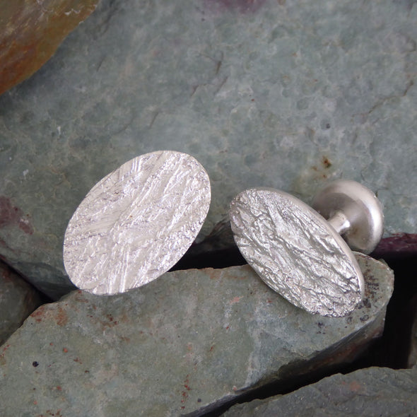 ein-paar-handgefertigte-manschetten-knoepfe-mit-gras-struktur-aus-sterling-silber-sie-bestehen-aus-einem-ovalen-und-einem-runden-plaettchen-die-mit-einem-festen-runden-steg-verbunden-sind-das-kleinere-runde-plaettchen-wird-wie-ein-knopf-durch-die-loecher-der-manschetten-gezogen-alexandra-baum-schmuck-frankfurt